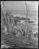 Man sitting on a tree with the ocean in the background, circa 1932-1939