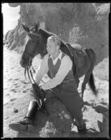 Gene Raymond, actor, sitting on a rock and holding the reins of a horse, circa 1933