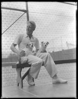Gene Raymond, actor, sitting on a bench next to a tennis court with two dogs, circa 1933