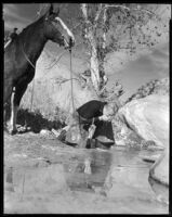 Gene Raymond, actor, drinking from a stream while a horse stands next to him, circa 1933
