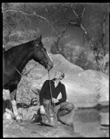 Gene Raymond, actor, kneeling by a stream and holding the reins of a horse, circa 1933
