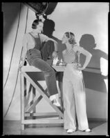 Woman sitting on a wooden platform while Allyn Drake, actress, stands below her, 1934