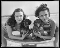 Geneva Mitchell and Mary Jo Mathews, actresses, on a beach, leaning on a table holding their pet dachsunds, 1934