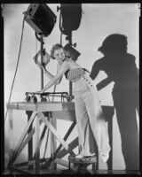 Allyn Drake, actress, standing on a stool and leaning on a spotlight that is set on top of a wooden platform, 1934