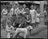 George Sidney and Charlie Murray, actors, sitting on a street corner surrounded by children and dogs, circa 1930-1934