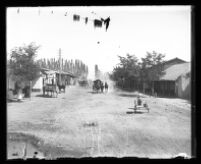 On unpaved street lined with small buildings, a horse and carriage approaches.